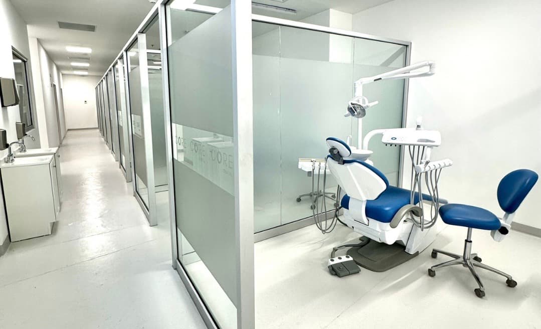 Consultorio dental con sillón dental azul, lámpara y bandeja de instrumentos detrás de una mampara de cristal