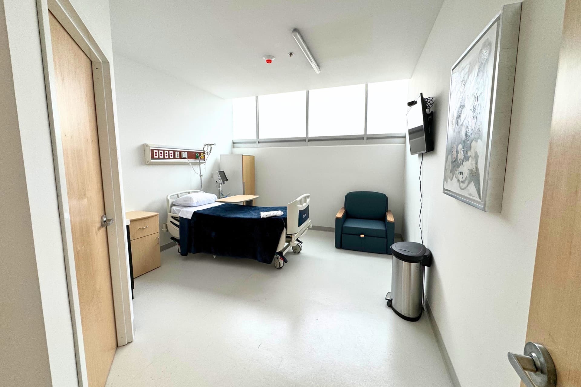 Habitación hospitalaria privada con cama, sillón reclinable, ventana y un cuadro en la pared