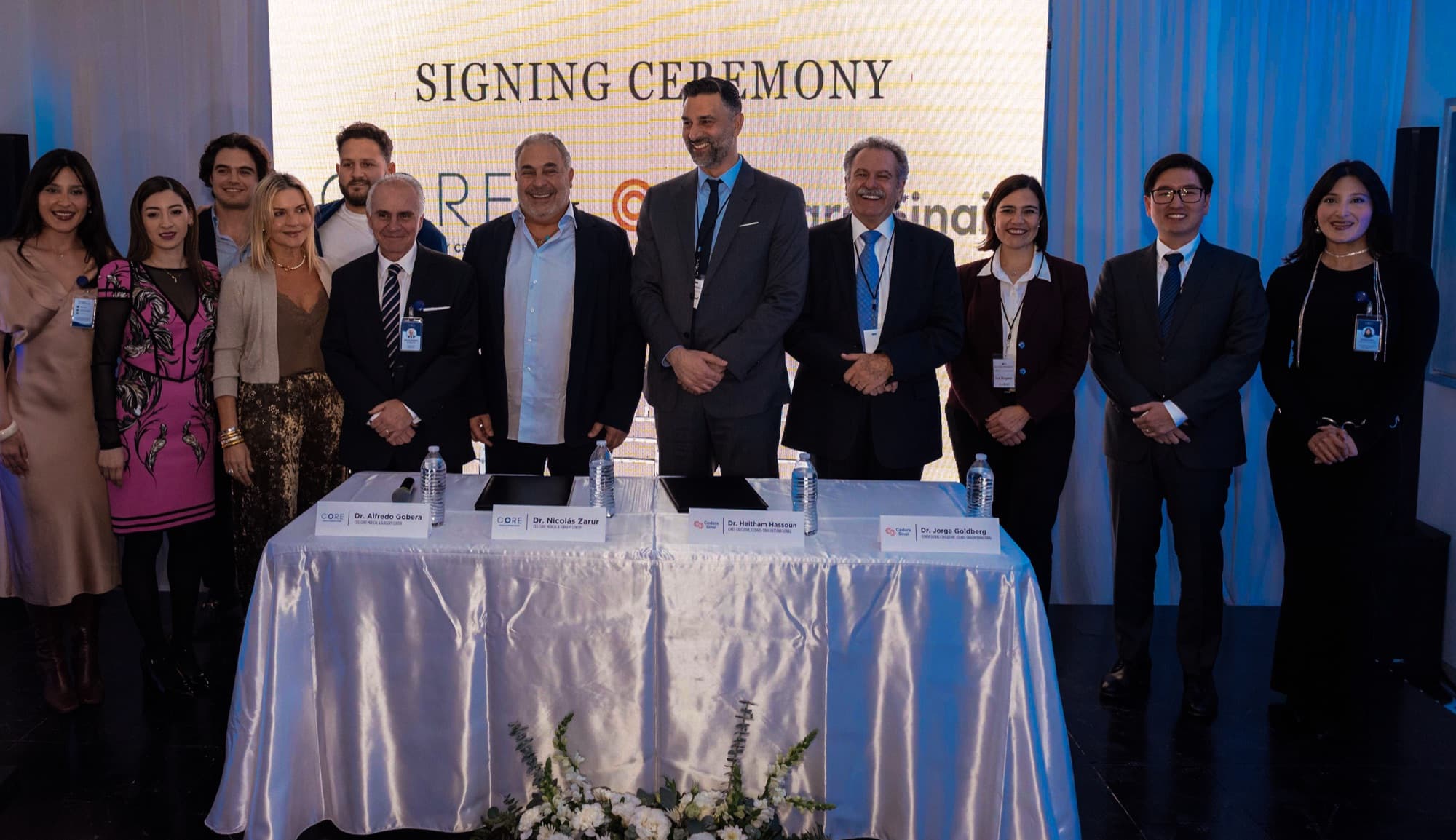 Eleven people stand in a row behind a draped signing table beneath a signing ceremony backdrop