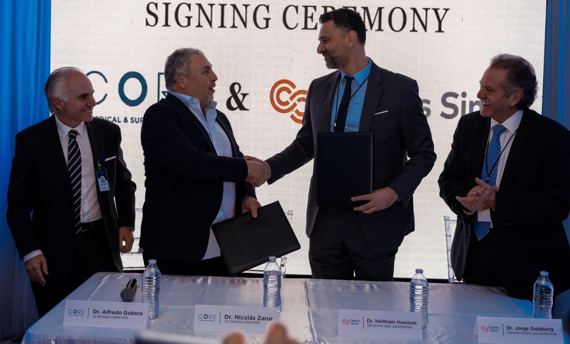 Two men shake hands across a signing table while two other men applaud, under a signing ceremony backdrop