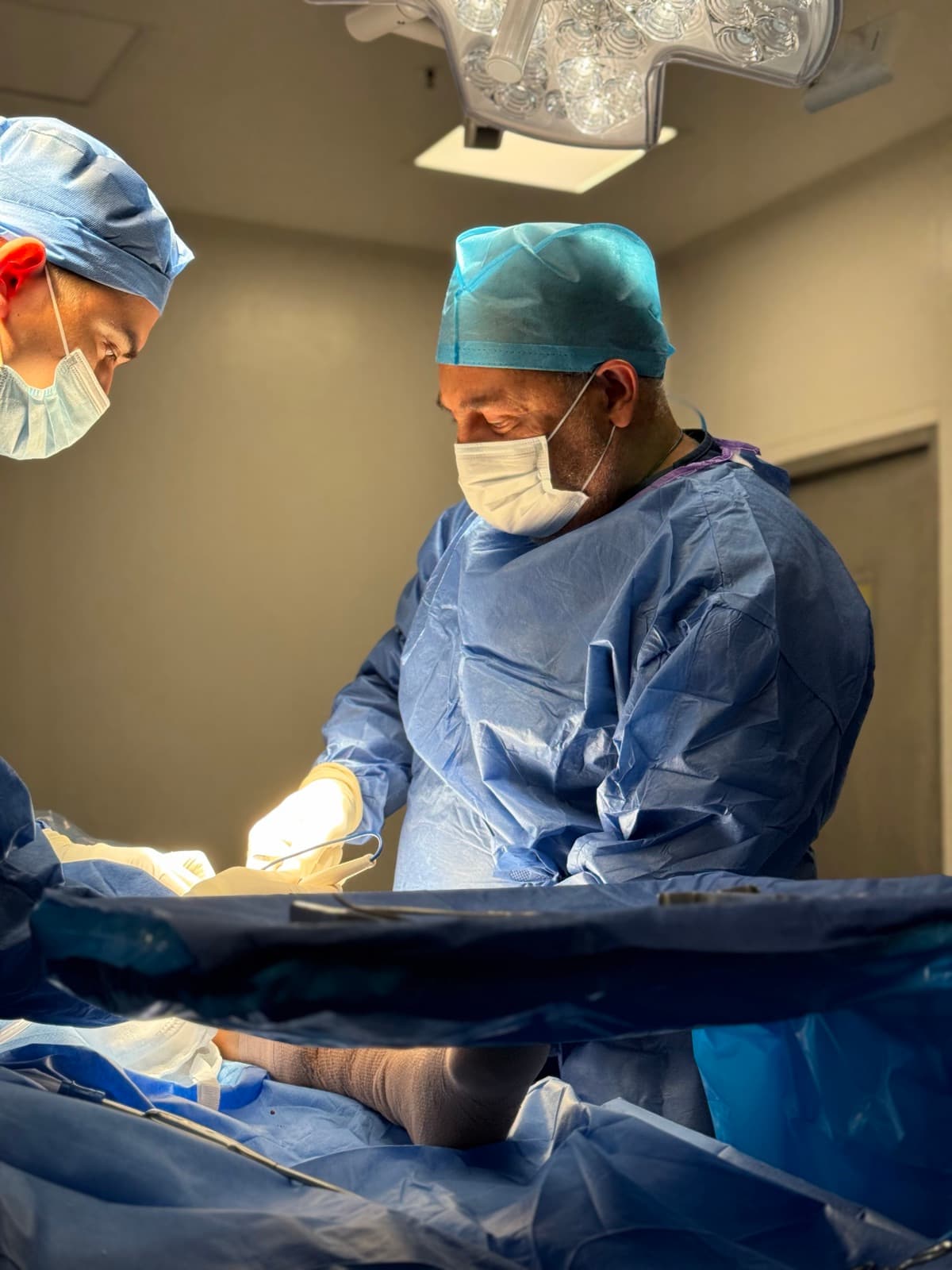 Two surgeons in sterile gowns operate at a draped table under surgical lights